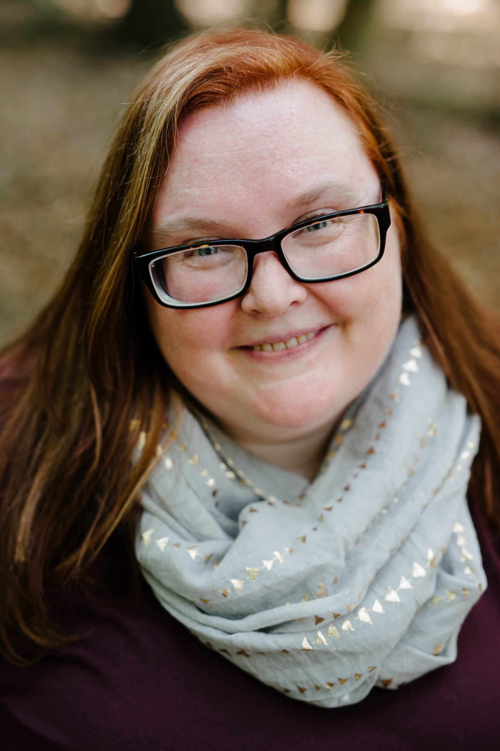 Headshot of a smiling white woman with long red hair, wearing glasses and a grey scarf around her neck.