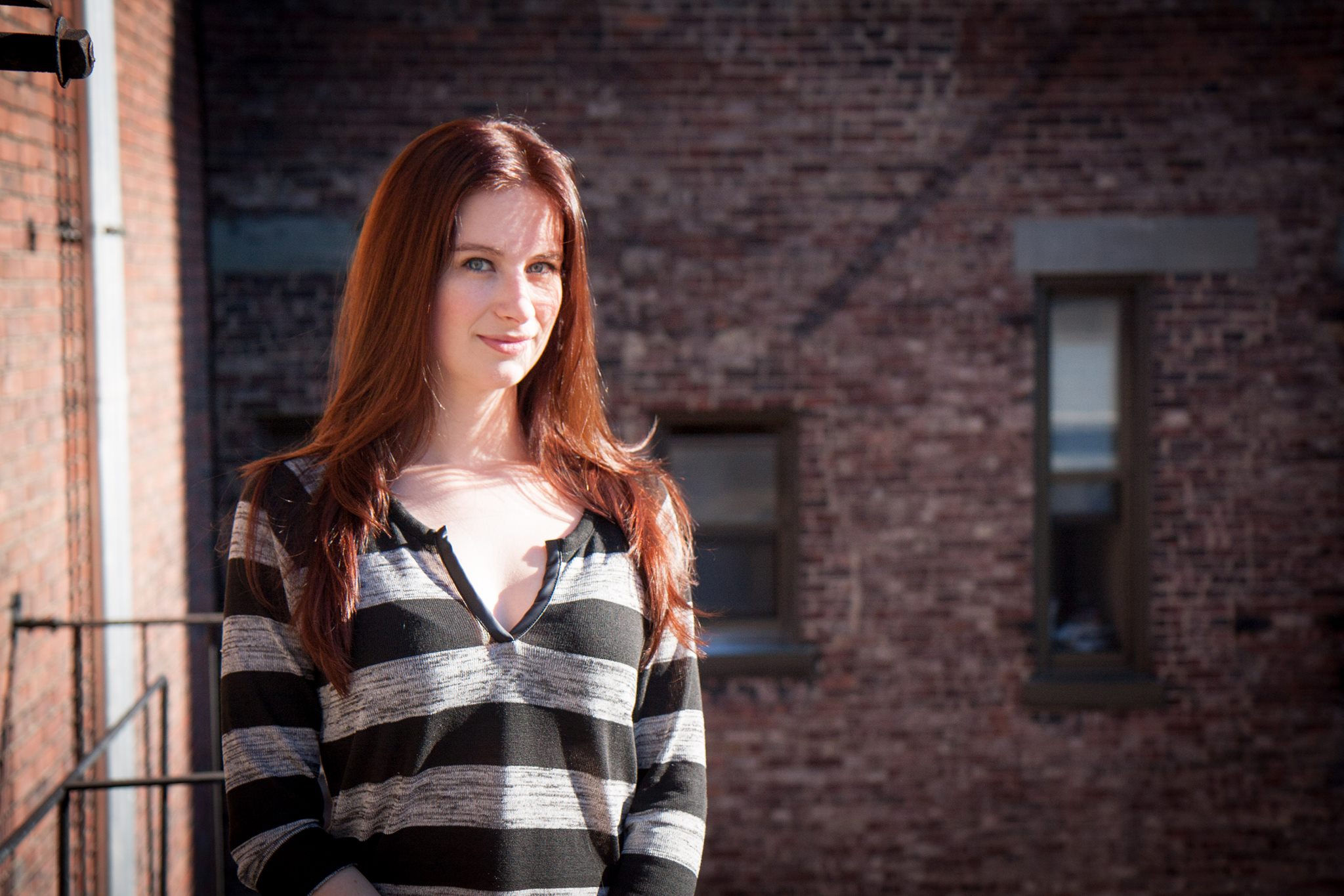 A white woman with long red hair wearing a grey and black striped shirt.